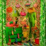 Highly patterned image of a standing figure with a purse over their head. They are holding several bags, and are surrounded by shoes. There are hats and bags on the wall behind them. A green dresser to the left had high heels coming out of the drawers. on March 12, 2025